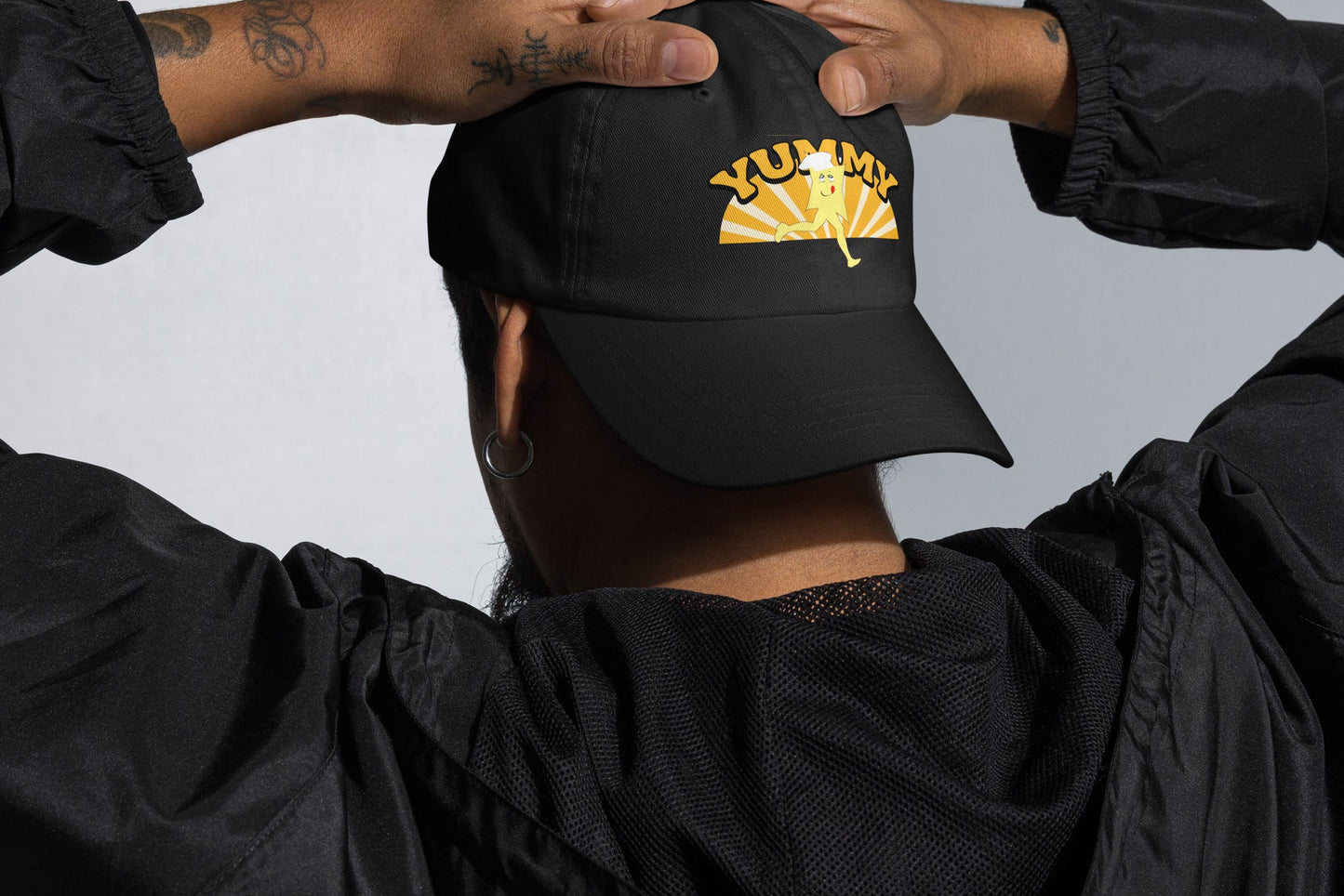 Person wearing a black cap with YUMMY character, hands clasped behind their head against a neutral background