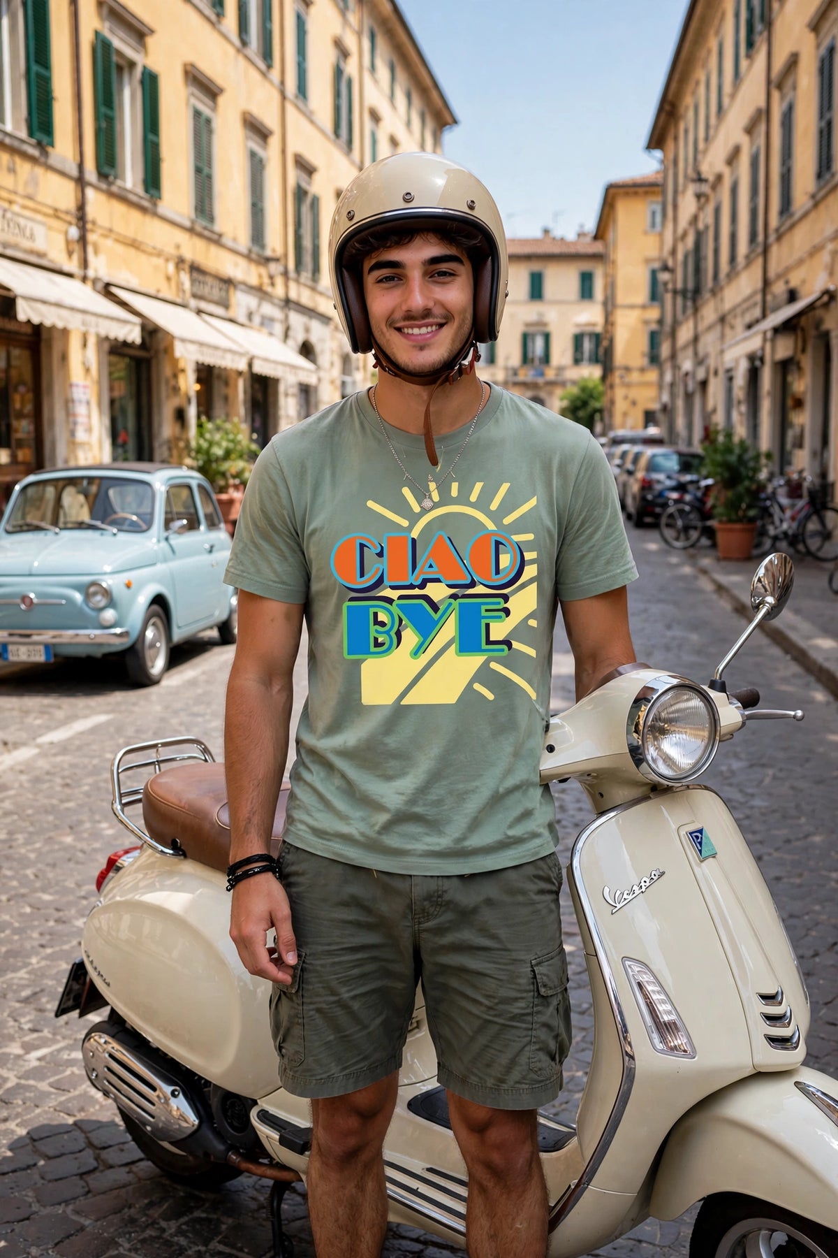 A man wearing a beige helmet and a green graphic t-shirt with Ciao Bye text stands next to a cream-colored Vespa scooter on a cobblestone street lined with yellow buildings.